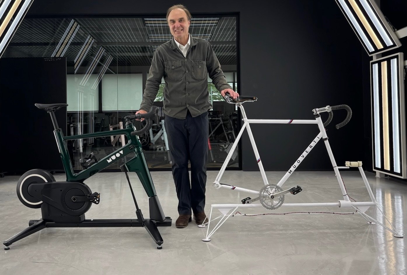 Alex Caccia standing between a dark green MUOV bike and the original white 2013 prototype in a MUOV studio