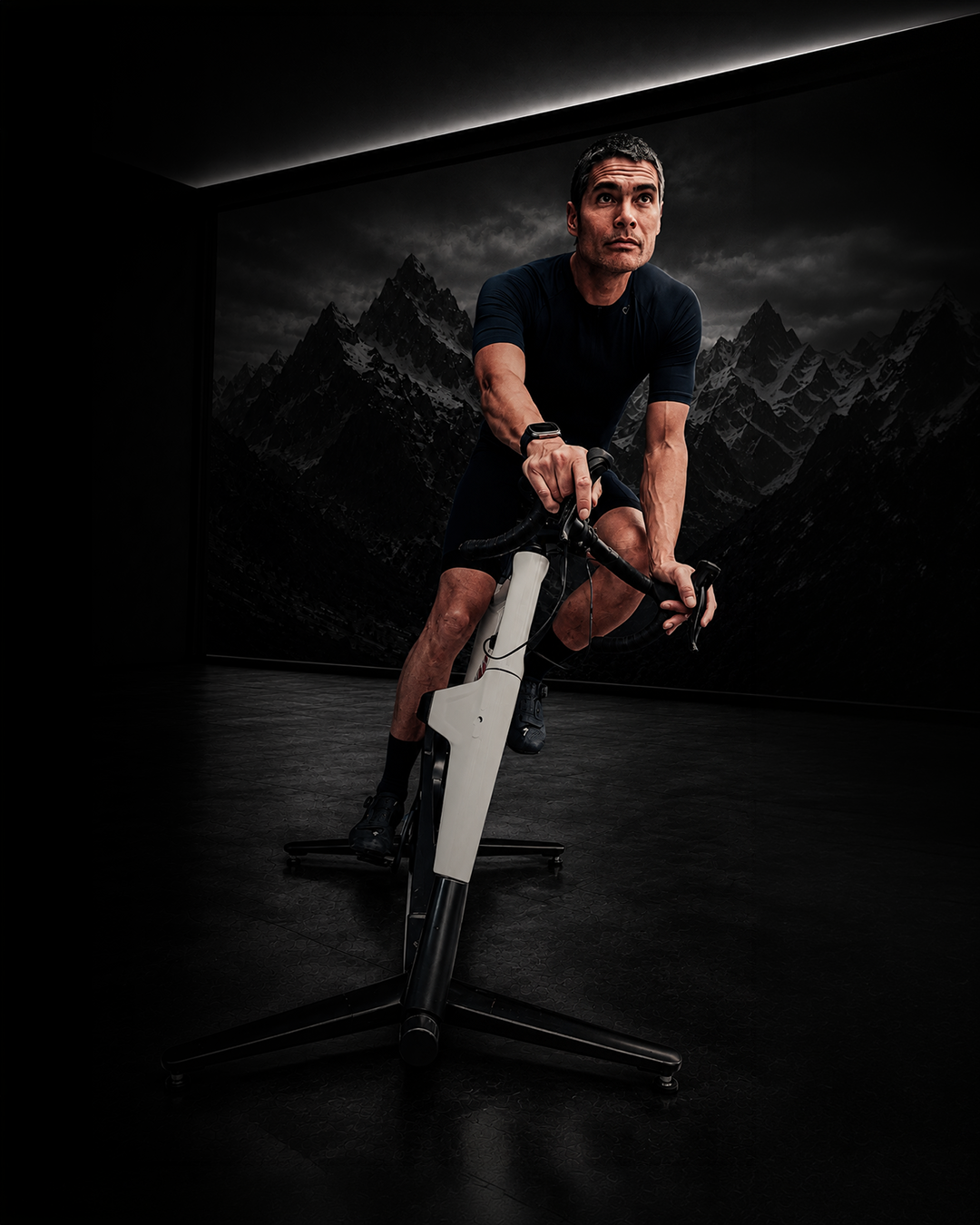 Rider leaning forward on a white MUOV bike in a darkened studio, a monochrome mountain backdrop behind